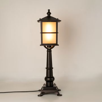 A black outdoor pillar lamp shown with light on against a white background highlighting its warm glow frosted panels and decorative metal pillar structure.