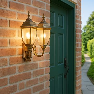 Double brass outdoor wall lantern with lights on mounted on a brick wall creating warm ambient glow with clear glass shades for elegant entrance lighting.