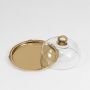 Brass serving cloche with clear glass dome shown on a white background highlighting its round tray and ornate handle with a clean minimal presentation.