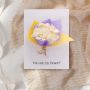 Greeting card with ivory paper flower bouquet wrapped in purple and yellow paper displayed on white background.