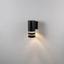 Minimalist black wall light on white background featuring a compact cylindrical body with dual rings.