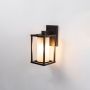 Outdoor wall light with black frame and frosted glass cylinder shade switched on against plain white background.