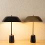 Two modern metal table lamps with wide dome shades in black and brass on stands with dark bases placed on a light surface against a textured wall.