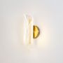Gold wall light with sculpted clear glass shade illuminated on a clean white background showing warm glow.