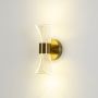 Gold wall light with sculpted clear glass shade illuminated on a clean white background showing warm soft glow.