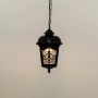 A classic black hanging lantern with the light on displayed against a white background highlighting its ornate design and softly glowing patterned glass panels.