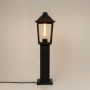 A tall black outdoor post light turned on displayed against a white background showing its sleek frame and clear glass panels with a glowing filament tube inside.
