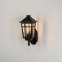 A black outdoor wall lantern with clear ribbed glass shown with light on against a white background highlighting warm illumination and structured lantern detailing.