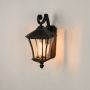 A black outdoor wall lantern displayed on a white background with the light on showing its clear patterned glass panels curved decorative arm and classic tapered roof design.